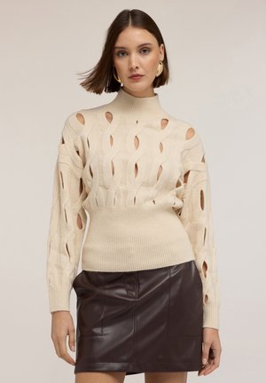 Beżowy sweter dzianinowy z wycięciami, wysoka szyja, ściągacze i dopasowana talia, w połączeniu z ciemnobrązową skórzaną mini spódnicą.