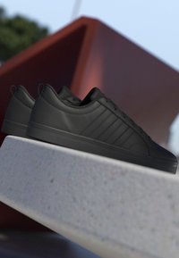 Paire de baskets basses noires avec des rayures texturées présentée sur une surface en pierre claire sur fond géométrique flou.