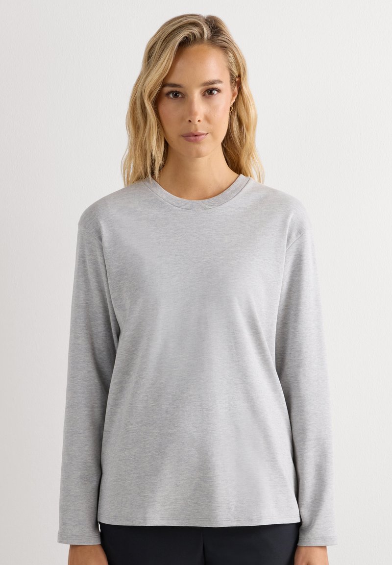 Jonge vrouw met lang blond haar die een effen lichtgrijs shirt met lange mouwen en ronde hals draagt, staand tegen een witte achtergrond.