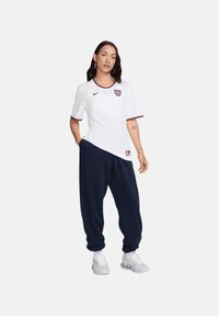 T-shirt sportivo bianco con dettagli rossi, dotato di un logo sul petto; abbinato a pantaloni della tuta blu navy e scarpe da ginnastica grigie.