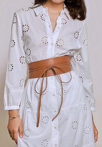 Witte hemdjurk met geborduurde bloemige uitsparingen, gecombineerd met een brede bruine leren riem met een strikdetail.