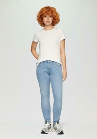 T-shirt blanche à manches courtes associée à un jean slim bleu clair et des baskets argentées. Le modèle se tient avec une main sur la hanche devant un fond neutre.