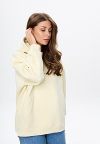 Helles gelbes übergroßes Hoodie aus weichem Material, mit einer Fronttasche, gerippten Bündchen und einer großen Kapuze. Kombiniert mit blauen Jeans.
