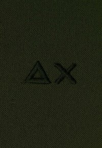 Tessuto verde scuro con una maglia testurizzata, caratterizzato da un logo ricamato a forma di triangolo e a forma di X in un verde leggermente più scuro.