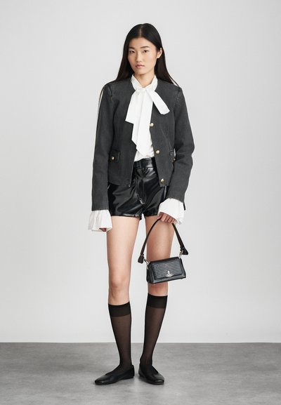 Jaqueta de denim preta com botões dourados, blusa branca com laço, calções pretos brilhantes, sapatos planos pretos e meias altas pretas até ao joelho. Bolsa com estampado de crocodilo.