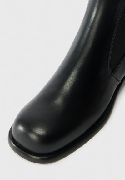 Czarne skórzane botki na kostkę o gładkiej teksturze, zaokrąglonym nosku i elastycznych panelach bocznych. Podeszwa jest płaska i nieco wyższa w pięcie.