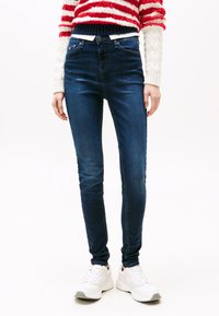 Plonos mėlyno džinso liesi džinsai su aukštu liemeniu, pasižymintys subtiliu išblukimu. Derinami su baltais sportiniais bateliais. Fonas yra baltas, vienspalvis.