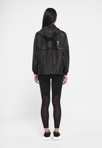 Kobieta w czarnej kurtce przeciwwiatrowej z kapturem, czarnych legginsach z przezroczystymi panelami i czarno-białych sneakersach, stojąca plecami.