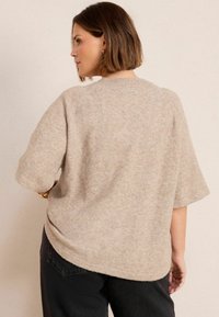 Beiger übergroßer Pullover mit kurzen Ärmeln, Rundhalsausschnitt und einem strukturierten, weichen Stoff. Der untere Saum ist leicht gewellt. Trägt schwarze Hosen.