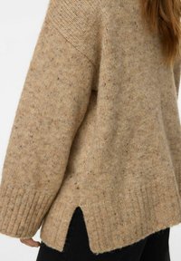 Jersey de punto beige con un acabado texturizado, con dobladillo acanalado y aberturas en los laterales para un detalle adicional, combinado con pantalones negros.