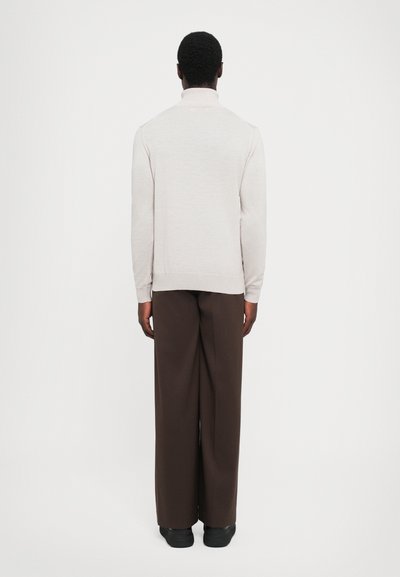 Maglione grigio chiaro a maglia con colletto alto, abbinato a pantaloni marroni larghi. L'outfit presenta una texture liscia e una vestibilità comoda.