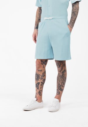 Hombre con pantalones cortos azul claro y zapatillas blancas, con piernas y brazos tatuados, de pie contra un fondo blanco.