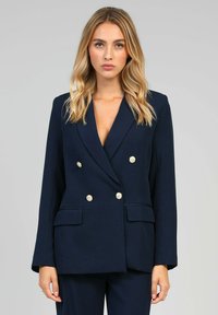 Blazer bleu marine à double boutonnage avec une coupe ajustée, boutons dorés, deux poches avant et revers cranté. Tissu lisse avec une forme structurée.