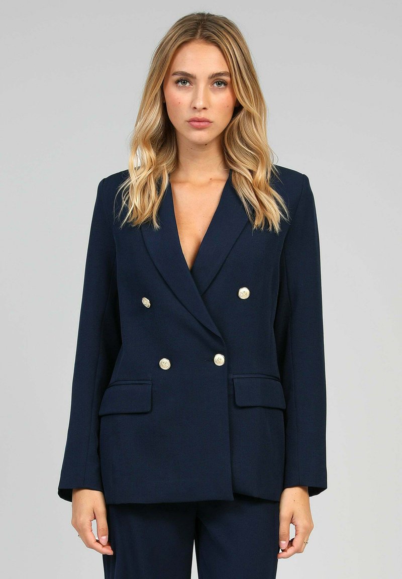 Blazer bleu marine à double boutonnage avec une coupe ajustée, boutons dorés, deux poches avant et revers cranté. Tissu lisse avec une forme structurée.