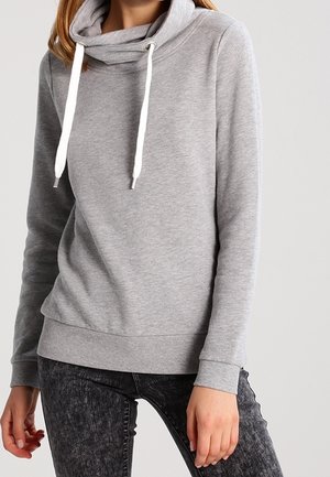 Femme portant un sweat-shirt à col roulé gris clair avec des cordons blancs et un jean gris foncé délavé, debout devant un fond uni.