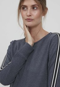 Felpa grigia con polsini a costine e colletto rotondo. Presenta dettagli a strisce bianche e nere lungo le maniche. Tessuto morbido.