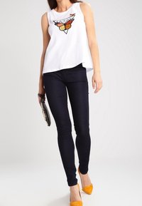 Top blanco sin mangas con un gráfico de mariposa, combinado con jeans oscuros ajustados y tacones amarillos de punta. Bolsa de mano en la mano.