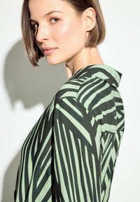 Blusa a righe nere e verdi con design con colletto. Presenta una texture morbida e una tasca sul lato. Lo sfondo chiaro esalta il motivo.