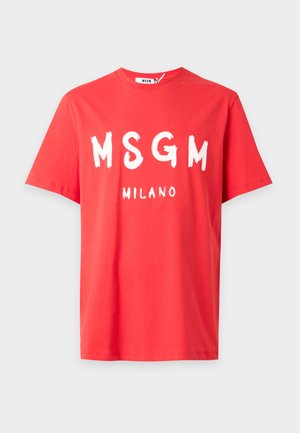 Мъж, носещ ярко червена тениска MSGM Milano, износени широки сини дънки и тъмни маратонки с червени връзки, стоящ с едната ръка в джоба.