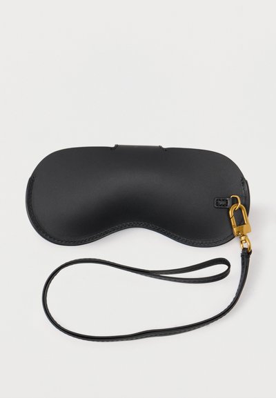 Μάσκα ύπνου από μαύρο δέρμα με καμπυλωτό σχήμα και προσαρμοσμένο μαύρο λουράκι με κούμπωμα σε χρυσό τόνο, πάνω σε λευκό φόντο.