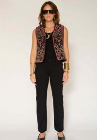 Gilet sans manches noir avec une broderie multicolore complexe, porté sur un haut noir et associé à un pantalon noir et des ballerines. Les accessoires comprennent des lunettes de soleil et des bracelets.