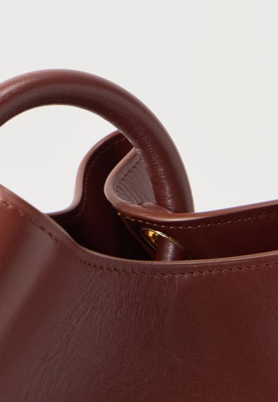 Κοντινό πλάνο σε καφέ δερμάτινη τσάντα με στρογγυλεμένες λαβές και ορατές ραφές σε ανοιχτό φόντο.