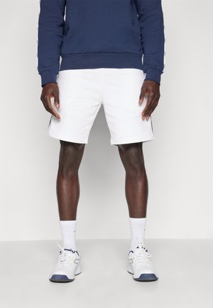 Mann, der weiße sportliche Shorts, weiße Sportsocken, weiße Turnschuhe und ein dunkelblaues langärmliges Sweatshirt trägt, steht vor einem schlichten Hintergrund.