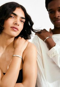 Twee personen dragen zilveren armbanden: één met een gehamerde textuur, de ander met een gladde afwerking. De achtergrond is wit, de kleding is minimalistisch.