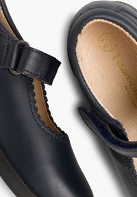 Zapatos infantiles de cuero negro con bordes ondulados, puntera redonda y una correa de velcro. El interior es de cuero beige con una marca de tamaño dorada.