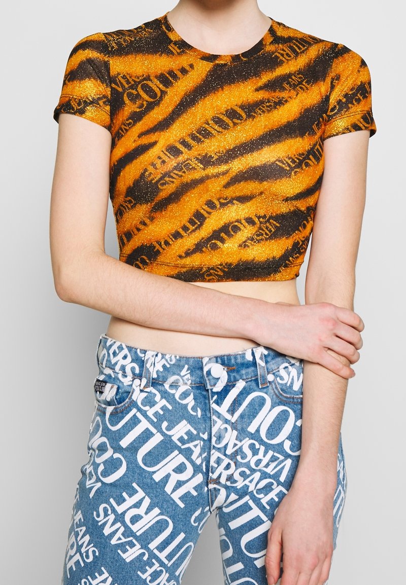 Pomarańczowo-czarny krótki top w paski tygrysie z brokatem, zestawiony z niebieskimi, wysokim stanem, jeansami ozdobionymi widocznymi białymi napisami.