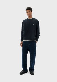Sudadera de corte marinero con puños de canalé, superpuesta a una camiseta gris, combinada con vaqueros de pierna ancha azules y zapatos negros.