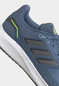 Chaussure de course Adidas bleue avec empeigne en mesh, bandes noires, accents vert néon et semelle blanche texturée vue de profil.