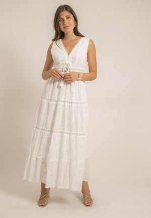 Femme debout portant une robe longue en dentelle blanche sans manches avec un décolleté en V et un détail noué, des talons beige à fines lanières, des boucles d'oreilles dorées et un bracelet.