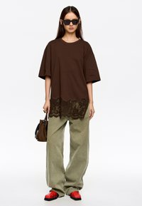 Brązowy, oversize t-shirt z koronkowym wykończeniem u dołu, zestawiony z jasnoszarymi, szerokimi spodniami oraz czerwonymi butami z teksturą węża. Czarny zegarek i torebka.