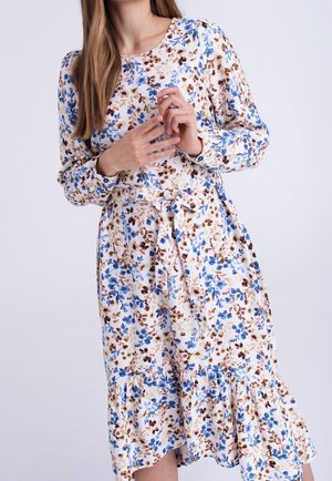 Kobieta ubrana w sukienkę w kwiaty z długim rękawem, z niebieskimi i brązowymi kwiatami, z wiązaniem w talii i falbaną na dole, stojąca na jednolitym tle.