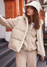 Gilet imbottito beige con un rivestimento morbido e soffice e pantaloni abbinati. Il modello indossa un cappello a secchiello beige, mettendo in risalto la palette di colori coordinata dell'outfit.