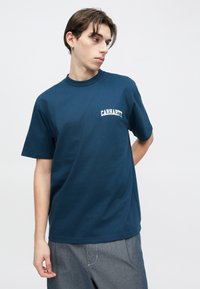 Μπλε ναυτικό βαμβακερό t-shirt με στρογγυλή λαιμόκοψη και κοντά μανίκια, με λευκά γράμματα "Carhartt" στην αριστερή πλευρά του στήθους.