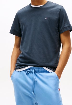 Mand iført en mørkeblå kortærmet t-shirt over en hvid skjorte med lyseblå sweatpants med snører, hænderne i lommerne.