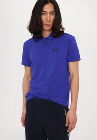 Polo da uomo in cotone blu brillante con una sottile trama. Presenta un colletto, maniche corte e un piccolo logo EA7 nero sul petto.