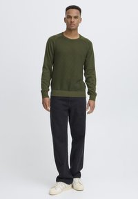 Suéter de punto texturizado en verde oscuro, con cuello redondo y puños acanalados, combinado con pantalones negros holgados y zapatillas blancas.