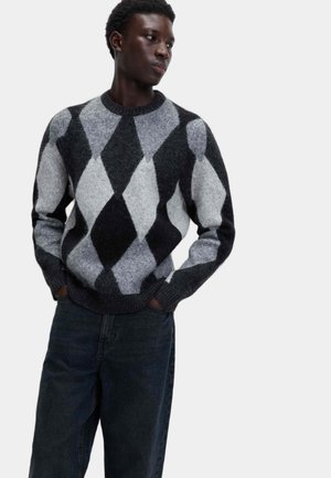Giovane uomo che indossa un maglione a rombi neri, grigi e bianchi, con jeans scuri, mani nelle tasche, che guarda di lato su uno sfondo bianco.