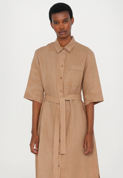 Vestido botonê cor castanho claro feito de um tecido texturizado, com mangas curtas, faixa na cintura, bolso no peito e bordado discreto do logo.