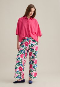 Chemise rose surdimensionnée avec col et fermeture à boutons, associée à un pantalon large à motif floral dans les teintes rose, vert et bleu. Chaussures plates marine.