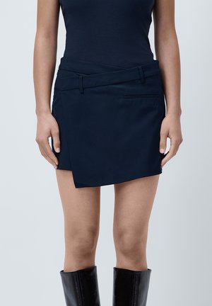 Navy blauwe wikkelrok gemaakt van een gladde stof, met een platte tailleband en een zijzak. Draagt een paar zwarte knie-hoge laarzen.