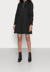 Černé denimové šaty s dlouhými rukávy, vysokým límcem a zapínáním na zip. Vybavené volánkovou sukní a hladkou texturou, doplněné černými kotníkovými botami.