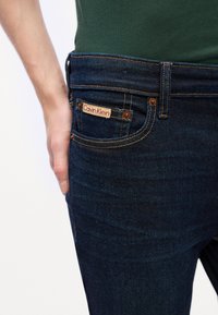 Κοντινή λήψη της τσέπης ενός σκούρου μπλε τζιν με ορατή ετικέτα Calvin Klein και ένα χέρι να ξεκουράζεται δίπλα στο ισχίο.