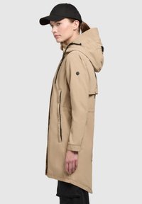 Parka impermeable beige con capucha, bolsillos con cremallera y detalles en negro. Presenta un corte relajado y una tela suave y ligera.