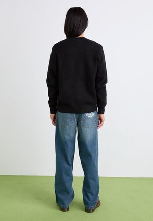 Person mit langen dunklen Haaren, die einen schwarzen Pullover und weite blaue Jeans mit sichtbarem weißem Logo auf der Gesäßtasche trägt, steht auf grünem Boden.