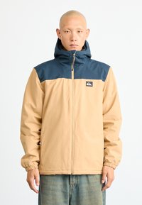 Man wearing beige and navy hooded jacket with Quiksilver logo, standing against plain white background.