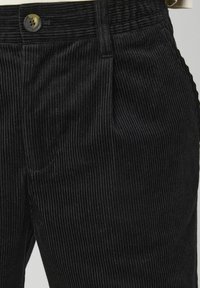 Pantalons en velours côtelé noirs avec une texture de côtes verticales, une fermeture à bouton et des poches latérales, partiellement visibles sur l'image.